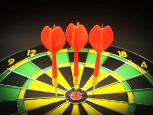 Three red darts are perfectly centered on the bullseye of a black, green, and yellow dartboard. The numbers 12, 18, and 4 are visible around the edge of the target.