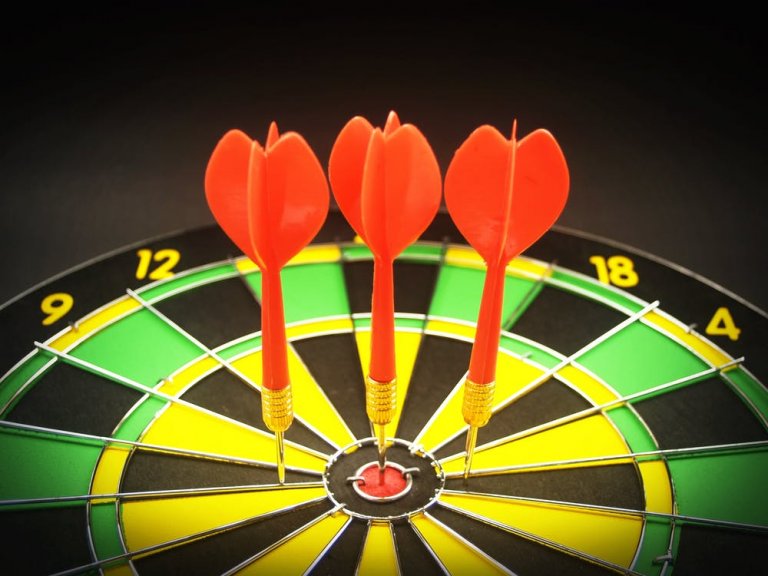 Three red darts are perfectly centered on the bullseye of a black, green, and yellow dartboard. The numbers 12, 18, and 4 are visible around the edge of the target.