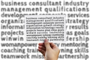 A hand holds a card filled with various business-related words, such as "business consultant," "qualifications," "development," "growth strategy," and "leadership." The background is blurred with similar target words.