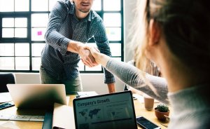 A man and a woman are shaking hands across a table in an office setting. The table has laptops, documents, and a small potted plant. A laptop screen facing the camera displays a presentation titled "Company's Growth," showing graphs and world maps highlighting target regions.