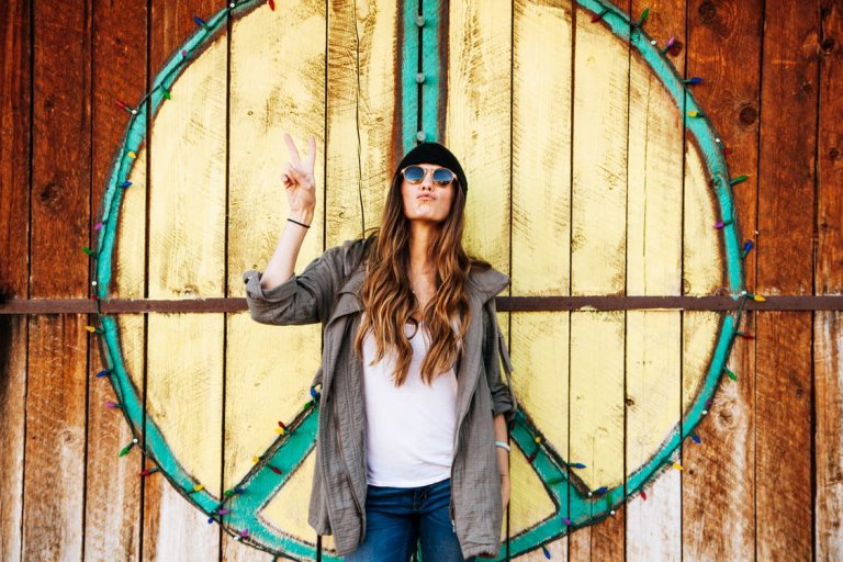 A person with long hair wearing sunglasses, a hat, a white shirt, and a gray jacket making a peace sign with one hand. They stand in front of a large peace symbol painted on a wooden wall decorated with colorful string lights, embodying the latest hiring trends in creative expression.