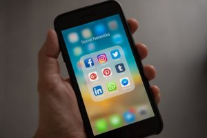 A person holds a smartphone displaying a folder labeled "Social Networks" with various social media app icons, including Facebook, Instagram, Twitter, Google+, Pinterest, Tumblr, LinkedIn, WhatsApp, and Messenger. The blurred background of other apps subtly hints at emerging hiring trends in the digital realm.