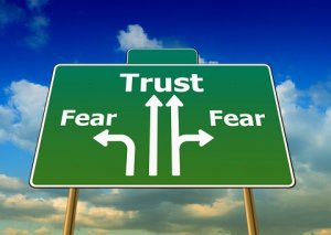 A green road sign with three white arrows pointing upwards. The central arrow, symbolizing trust, is labeled "Trust," while the left and right arrows are labeled "Fear." The sky in the background is blue with scattered clouds.