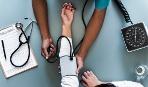 A healthcare professional measures a patient's blood pressure using a sphygmomanometer. The patient’s arm is extended on a table, striving to maintain their healthiest state, while medical tools including a stethoscope and a clipboard with papers are visible nearby.