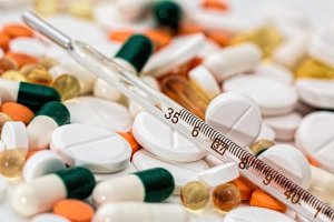A pile of various medications including white tablets, orange pills, green and white capsules, and oval gel capsules rests on the table. A thermometer displaying a temperature of approximately 38 degrees Celsius lies atop this assortment, a stark contrast to the healthiest state one aspires to achieve.