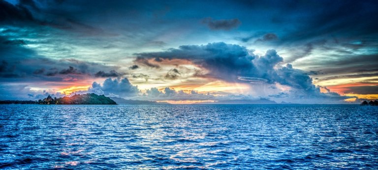 A striking holiday seascape at sunset with a vivid, colorful sky dominated by dramatic clouds. The horizon glows with hues of orange, yellow, and pink, reflecting on the calm, deep blue ocean. An island with silhouetted trees is seen on the left.