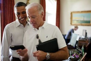 Two men, one in a white polo shirt with sunglasses on his collar and the other in a dark suit, stand close together in an office, smiling at a smartphone screen. The man in the polo holds a folder, possibly planning their next holiday adventure.