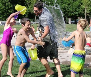 A group of children and an adult are having fun and laughing during a holiday, splashing each other with water in a park. The adult is getting sprayed with a large splash from a bucket, adding to the energetic and lively scene, with green trees in the background.