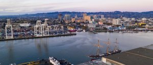 A panoramic view of a marina at dusk in Oakland, featuring a tall ship docked near the waterfront. The scene includes an array of cranes and containers, suggesting an industrial port area. The skyline of the city is visible in the background, with hills in the distance.
