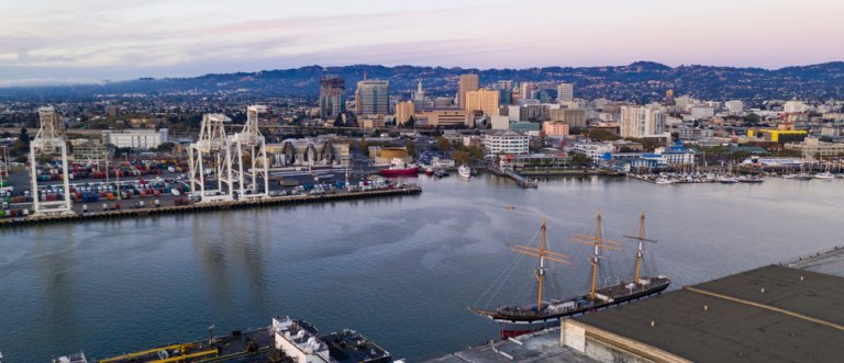 A panoramic view of a marina at dusk in Oakland, featuring a tall ship docked near the waterfront. The scene includes an array of cranes and containers, suggesting an industrial port area. The skyline of the city is visible in the background, with hills in the distance.