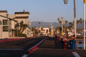 A quiet boardwalk lined with buildings, palm trees, and street lamps in warm sunlight, with mountains visible in the background and a clear blue sky overhead.