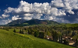 A scenic landscape featuring Concord’s lush, green valley with a small town nestled among trees and rolling hills. Majestic mountains rise in the background, topped with fluffy white clouds against a vibrant blue sky. Hints of sunlight create a serene and picturesque scene.
