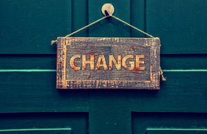 A weathered wooden sign with the word "CHANGE" hangs from a rope on a dark green door. The sign's rustic appearance contrasts with the smooth, painted surface of the door, emphasizing the message of transformation—a testament to hard work and renewal.