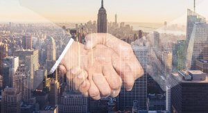 A double exposure image featuring a close-up of two hands shaking in the foreground overlaid on a cityscape view of tall skyscrapers, including the Empire State Building, at sunset. The handshake suggests a successful business deal or partnership with implications involving the federal government.