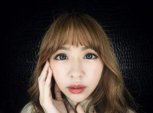 A close-up of a millennial with long, light brown hair and bangs, posing against a dark, textured background. The person has expressive, wide eyes and is gently touching their face with both hands. They have a neutral expression.