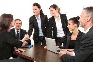 A group of six business professionals, three women and three men, dressed in formal attire, are meeting in a conference room. Two millennial women are standing and smiling, while the other four are seated at a table with a laptop. One woman is shaking hands with a man.