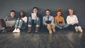 Six millennials sit on the floor in a row, casually dressed and smiling. They hold various electronic devices including a laptop, smartphone, and tablets. The background is a plain, dark wall, and the wooden floor adds warmth to their relaxed and happy atmosphere.