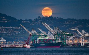 A large cargo ship is docked at the Oakland port, with multiple cranes surrounding it. In the background, a large orange full moon rises over a hilly area, creating a dramatic evening scene. The ship is prominently labeled "EVERGREEN.