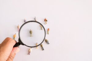 A recruiter's hand holds a magnifying glass over a group of miniature figurines standing in a circle on a white background. The magnifying glass highlights one figurine standing in the center, emphasizing its details and significance.