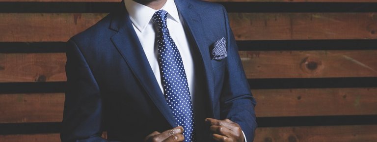 A person in a navy blue suit and white shirt adjusts their suit jacket with confidence, exuding self-esteem. They wear a polka dot tie and matching pocket square. The background features horizontal wooden slats. The image captures the upper torso, leaving the person's face unseen.