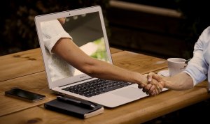 A person shakes hands with another person whose arm is extending out from a laptop screen, symbolizing a virtual handshake or online agreement. The background includes a wooden table with a smartphone, notebook, pen, and coffee cup, highlighting the modern touch of social media's influence on business deals.