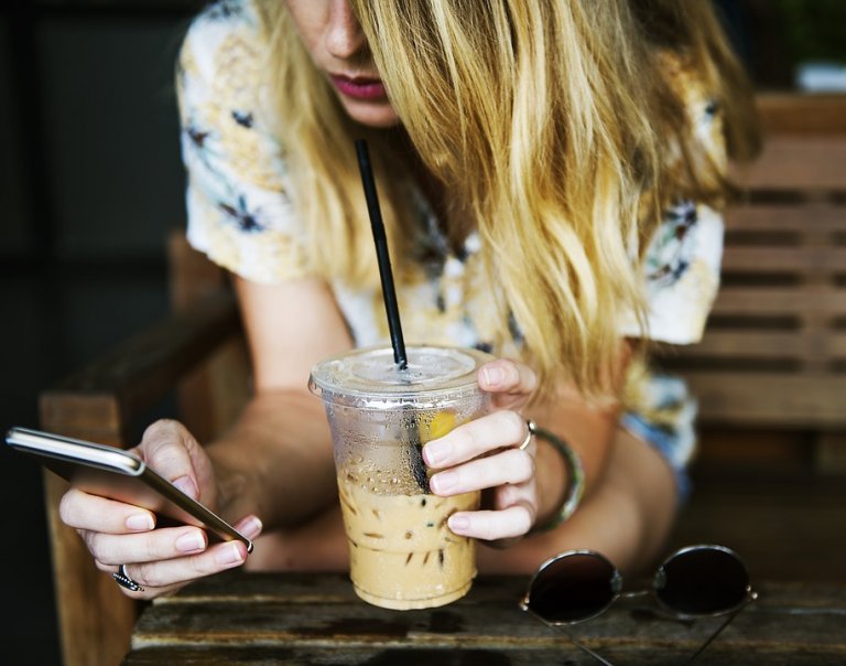 A person with long blonde hair is sitting and holding a plastic cup of iced coffee with a black straw in one hand while using a smartphone with the other. The person is wearing a floral shirt, and there are sunglasses on the table in front.