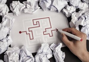 A hand holding a red pen is completing a maze on graph paper, symbolizing the challenges of employee engagement. The maze is surrounded by numerous crumpled sheets of paper, suggesting previous failed attempts. The partially drawn path leads from a starting point to an endpoint.