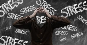 A person stands in front of a chalkboard with "STRESS" written multiple times on it. The person's head is replaced with the word "STRESS," and they hold their head in their hands, as if the temperature of their anxiety is on the rise, conveying a sense of overwhelming stress.