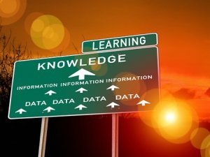 A green road sign against a sunset sky indicates "LEARNING" with an arrow pointing upwards towards "KNOWLEDGE". Below, smaller arrows point towards "INFORMATION" and "DATA", symbolizing the path from data to wisdom through learning.
