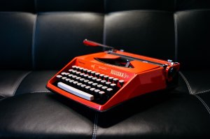 A vintage red typewriter with white keys and black lettering sits on a black leather surface, reminiscent of tools once used in a budding government career. The word "Riviera" is prominently displayed on the front. The lighting highlights its retro design and vibrant color.