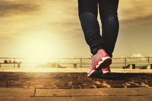 A person wearing jeans and pink athletic shoes is walking on a cobblestone path. The background features a sunset or sunrise by the water, with a railing and sky in warm tones, reflecting the calm one seeks after a busy day nurturing their government career.