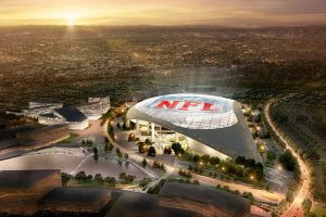 An aerial view of an expansive stadium lit up at dusk with "NFL" prominently displayed on the roof. The surrounding area includes modern buildings, parking lots, and lush landscaping. The cityscape extends into the distance under a glowing sunset, showcasing a perfect blend of building brand identity and urban development.