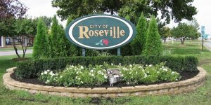 A landscaped sign that reads "City of Roseville" with a red rose graphic is nestled among green bushes and small white flowers. Situated within a circular stone planter, with trees and Concord Road visible in the background, it captures the community spirit perfectly.