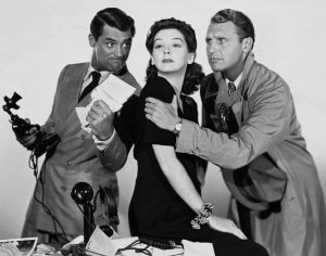 Three individuals in a black and white scene: a woman sits while two men flank her. The man on the left holds a telephone and papers, resembling a resume template, wearing a suit and smiling. The man on the right, in a trench coat, leans protectively toward the woman, who looks away thoughtfully.