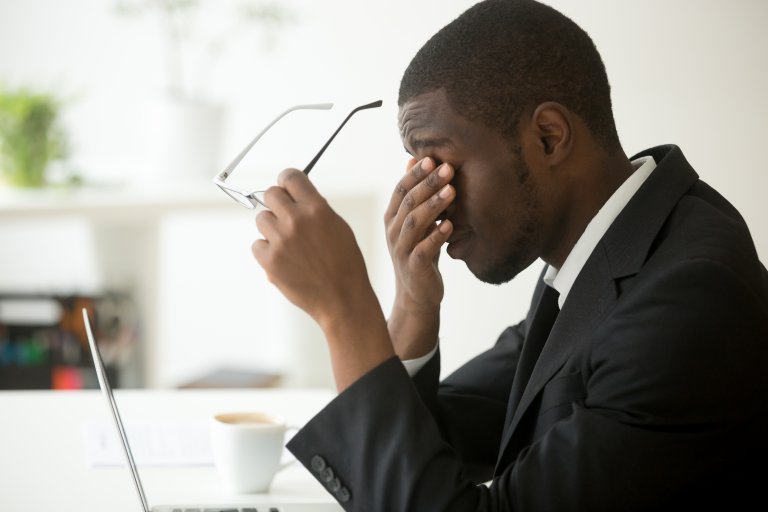 A man in a business suit sits at a desk with his laptop open. He holds his glasses in his right hand and covers his eyes with his left hand, appearing stressed or tired, perhaps reflecting on employee engagement challenges. A coffee cup and some papers are on the desk in the bright office space.