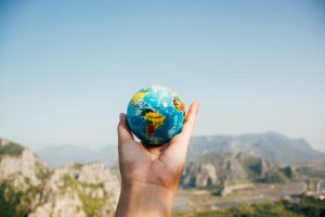 A hand holding a small globe against a scenic background with mountains and a bright blue sky, strategically showcasing countries and continents in vibrant colors.