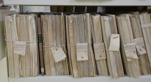 A close-up view of a shelf filled with old, worn files and documents tied together with string and labeled with handwritten tags. A single dark brown leather-bound book stands out strategically among the beige folders.