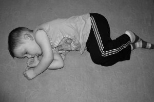 Black and white photo of a small child lying on their side on the floor, asleep. The child is wearing a short-sleeve shirt, dark pants with stripes on the side, and striped socks. They are curled up, with one arm under their head and the other resting near their mouth—a scene that evokes a sense of calm similar to true employee engagement.