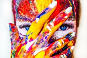 A close-up of a person with their face and hand covered in vibrant, multicolored paint. They have striking blue eyes peeking through the gaps between their fingers, which also have painted fingernails—a true portrait of creative success. The background is blurred and indistinct.