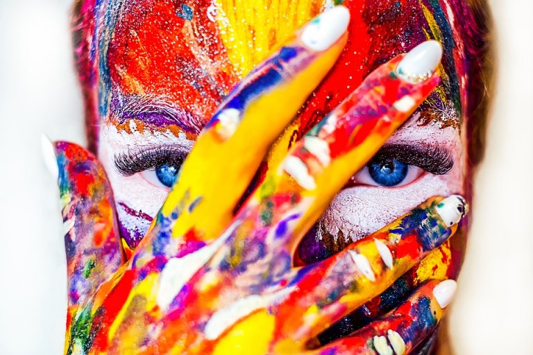 A close-up of a person with their face and hand covered in vibrant, multicolored paint. They have striking blue eyes peeking through the gaps between their fingers, which also have painted fingernails—a true portrait of creative success. The background is blurred and indistinct.