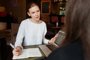 A woman with a focused expression is sitting at a table with a notebook and pen in hand, gesturing towards another person whose back is turned. She has a laptop open in front of her, suggesting an engaged discussion or a business meeting aimed at achieving success.