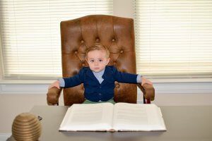 A young child sits in a large brown leather chair behind a desk, completing homework from an open book with visible text. Arms rest on the chair's armrests. The background features closed blinds covering windows. The child is dressed in a navy blue blazer and a light blue shirt.