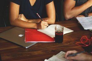 Close-up of three people sitting at a wooden table engaged in a homework session. One person is writing in a notebook with a red folder beside a laptop, another takes notes with a pencil, and a jar of dark liquid is on the table. Notebooks and papers are scattered around.