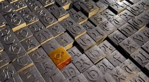A large number of dark metallic letter blocks are arranged in a grid pattern. Among them, one golden block stands out prominently, displaying the symbol for infinity. The blocks resemble an intricate cover letter with various letters and symbols etched into their surfaces, creating a textured effect.