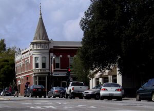 A street scene in Concord features a mix of cars stopped at a stoplight in front of a historic brick building with a turret. Trees line the sidewalk, and pedestrians are visible near the crosswalk. The sky is partly cloudy.