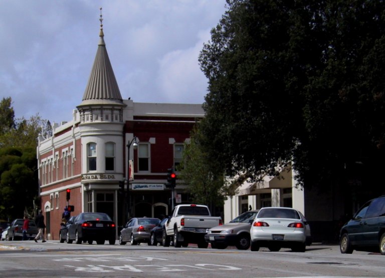 A street scene in Concord features a mix of cars stopped at a stoplight in front of a historic brick building with a turret. Trees line the sidewalk, and pedestrians are visible near the crosswalk. The sky is partly cloudy.