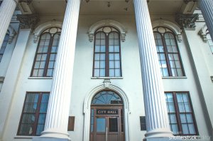 A grand building with tall white columns and large arched windows, featuring a central wooden door labeled "CITY HALL" in white letters. The facade is classical with ornate details, a symmetrical design, and a sense of historical significance that defines the city brand.