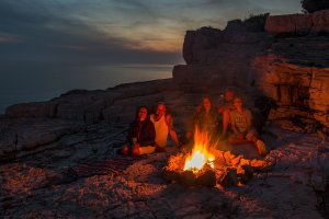 A group of people sitting around a campfire on a rocky shore at dusk, sharing stories. The sky is darkening with a beautiful sunset on the horizon, and the calm ocean is visible in the background.
