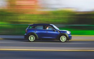 A blue compact car with green wheels is driving at high speed on a road with blurred greenery in the background. The driver, visible through the window, seems to be racing through countless stories as the motion blur emphasizes the vehicle's fast movement.