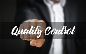 A person in a business suit points towards the viewer with their index finger, as if inviting them into the narrative of quality control. The words "Quality Control" are prominently displayed in white cursive over the image. The background is blurred, emphasizing both the text and the gesture, creating a compelling visual story.